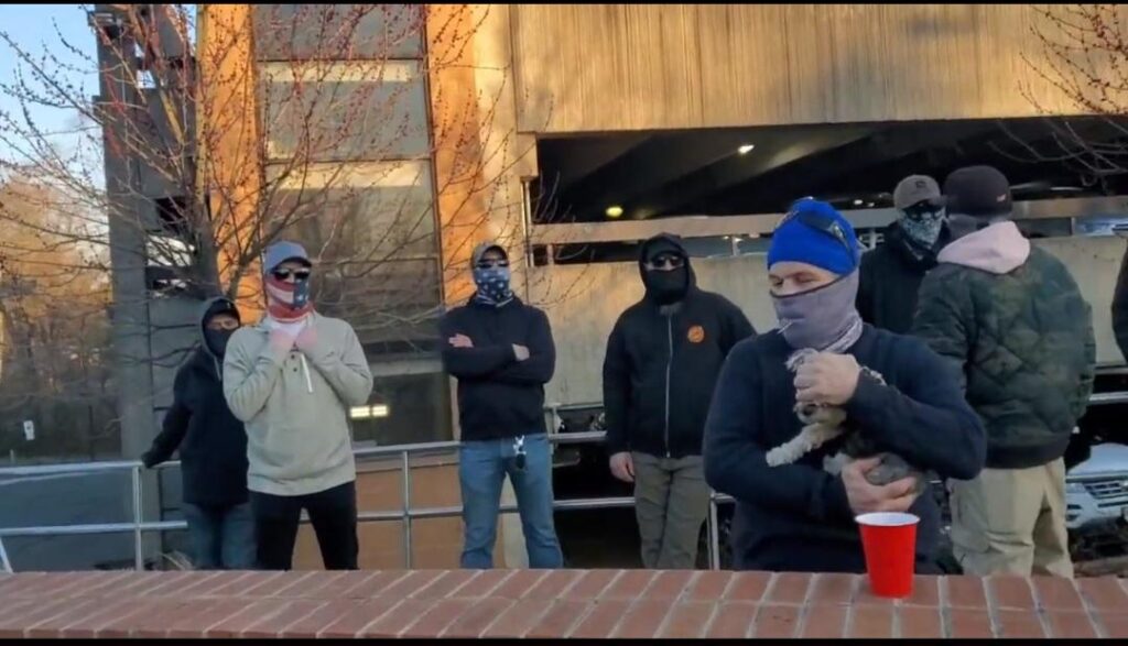 Seven masked individuals, one of them holding a dog, standing behind a brick embankment with a parking deck in the background.